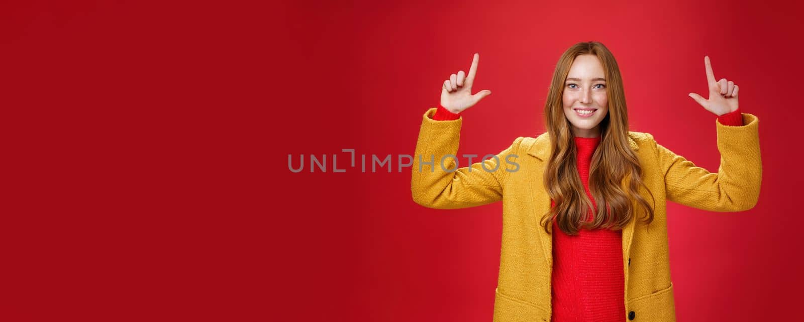 Indoor shot of redhead attractive woman in yellow fall coat raising hands promoting advertisement as pointing up and smiling broadly with satisfied pleasant expression over red background. Copy space