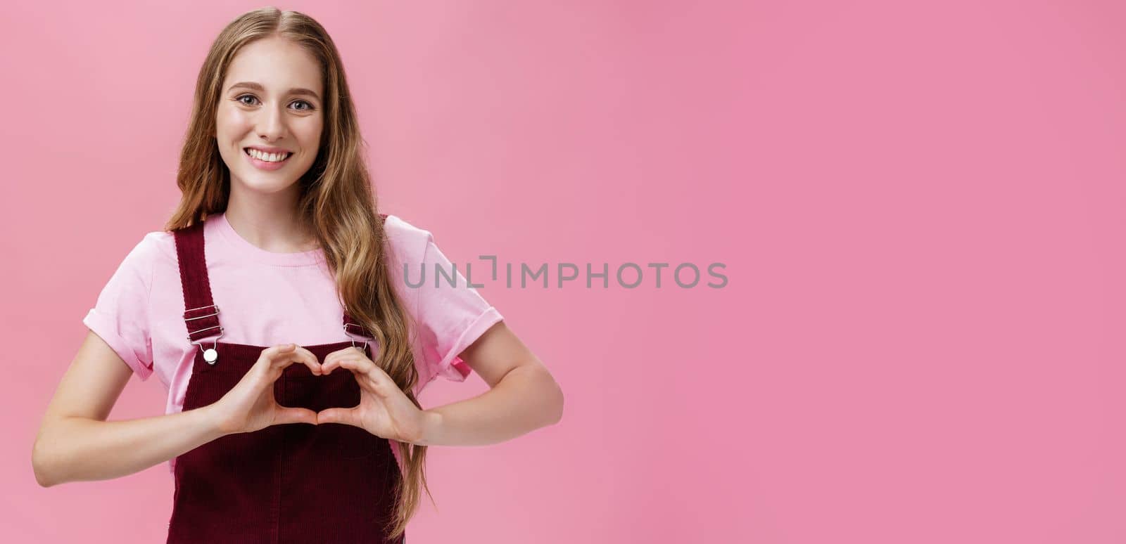 Girl loves family. Kind charming young woman in overalls with small tattoo on arm showing heart gesture over body and smiling lovely at camera expressing tender and cute attitude over pink wall by Benzoix