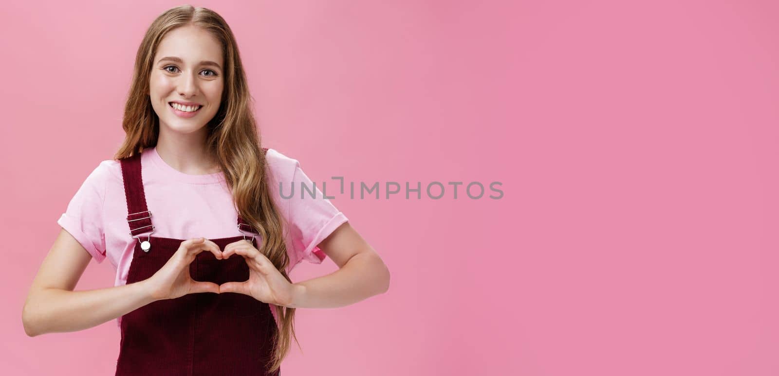 Girl loves family. Kind charming young woman in overalls with small tattoo on arm showing heart gesture over body and smiling lovely at camera expressing tender and cute attitude over pink wall.