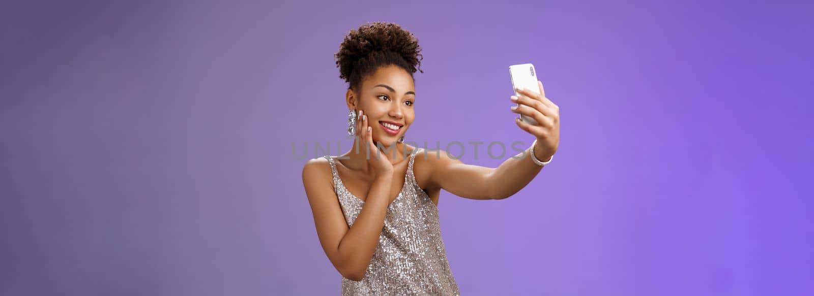 Popular stylish african-american female. blogger take selfie partying night club extend arm holding smartphone posing looking phone display gladly grinning standing blue background in elegant dress.