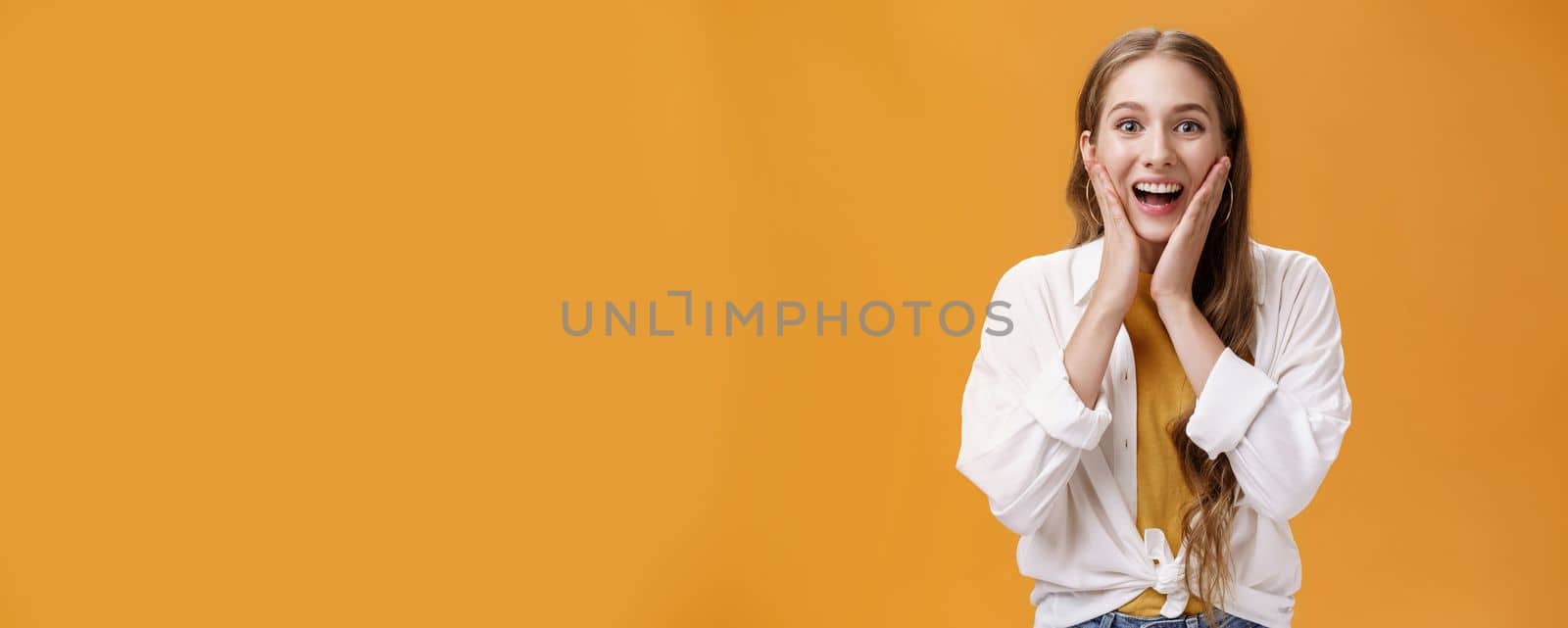 Excited and amused pretty young stylish woman with tattoo in trendy outfit touching cheeks from surprised and thrill gazing entertained at camera hearing awesome news, posing over orange wall. Body language concept