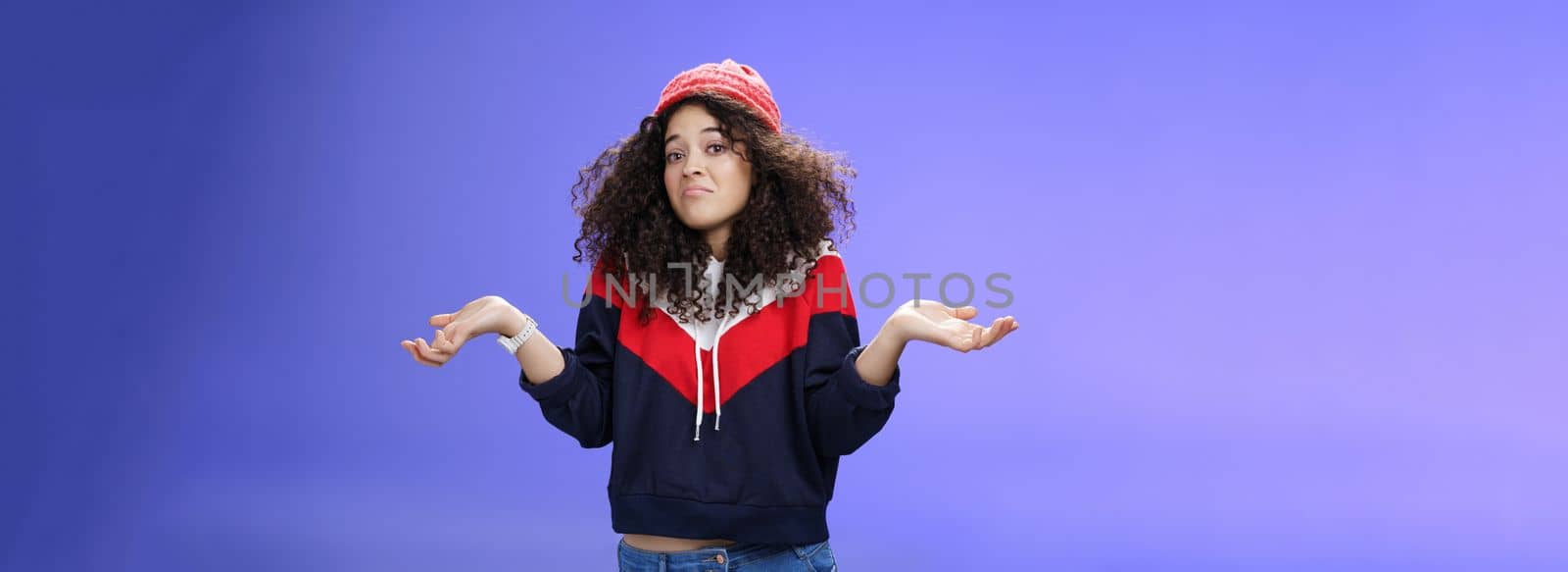 Upset girl cannot get clue what happened shrugging turning away as pouting feeling questioned and unsure what answer, standing clueless and puzzled in sweatshirt and stylish hat over blue background.