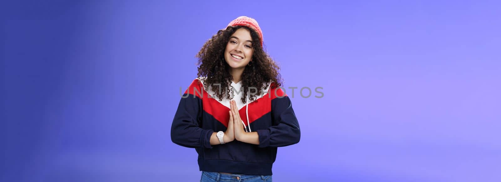 Portrait of charming curly-haired feminine girlfriend begging for favour as posing over blue background in winter outfit with hat, holding hands in pray smiling with angel look and friendly gaze. Body language and facial expressions concept