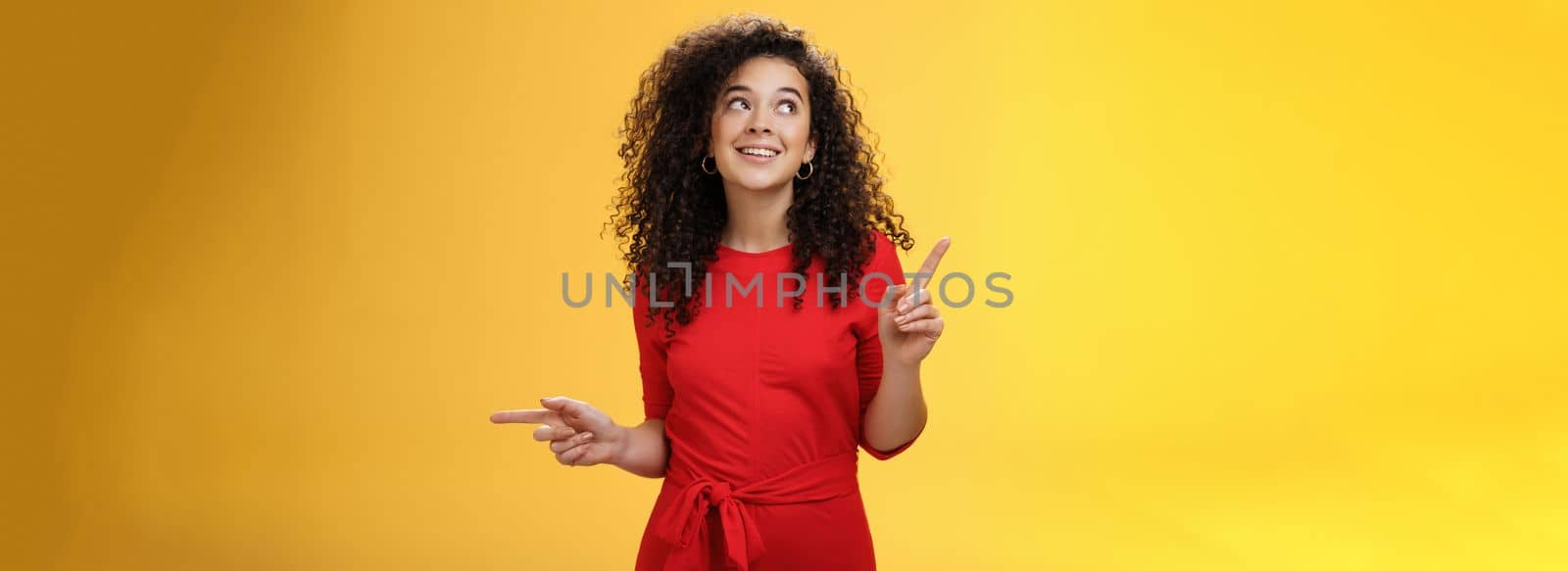 Dreamy charming woman making choice as checking out two awesome products pointing sideways looking intrigued and curious at upper left corner, smiling dreamy and delighted over yellow wall by Benzoix