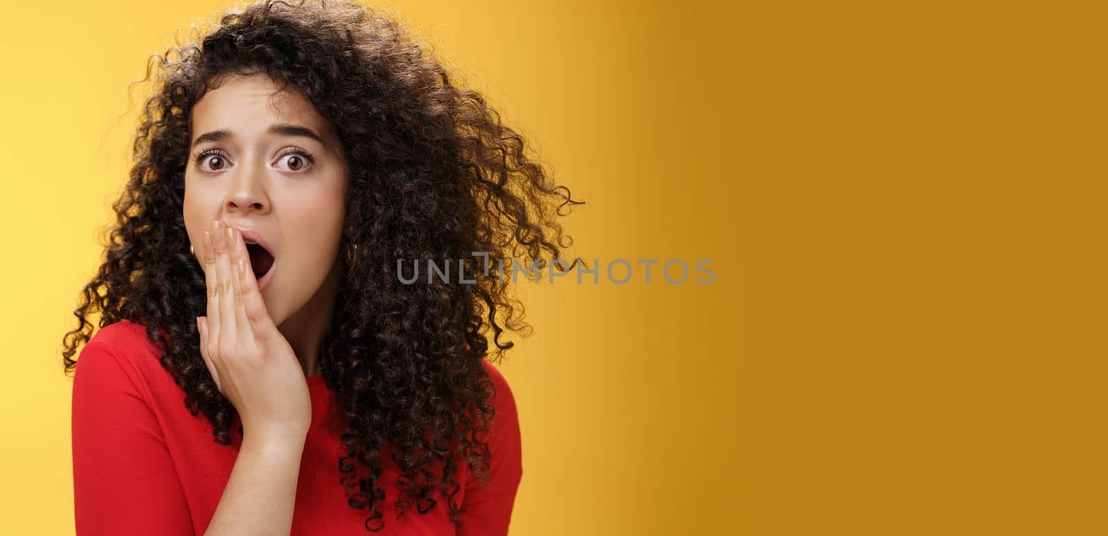 Close-up shot of concerned and shocked worried woman with curly hairstyle open mouth wide and covering it with palm popping eyes at camera impressed and upset over yellow background. Emotions and faciale xpressions concept