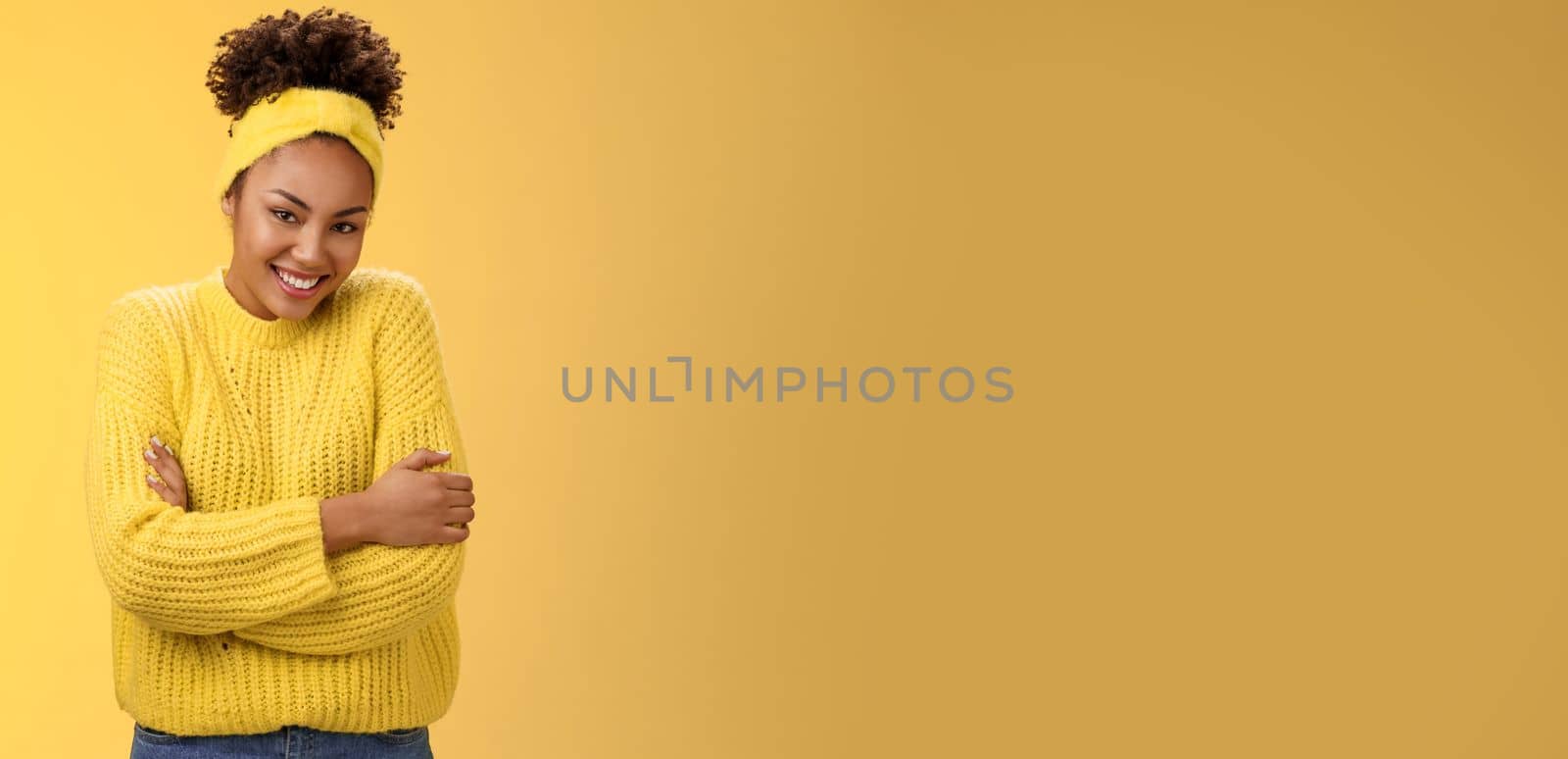 Tender delicate young african-american cheerful girlfriend curly hairstyle headband sweater hugging herself embracing gladly smiling camera feel soft comfortable, standing yellow background warm.