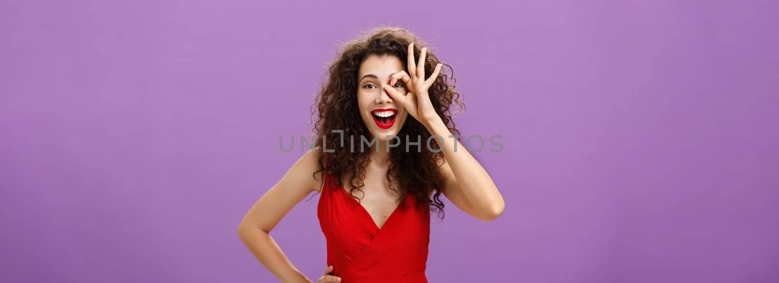 I got zero worries. Portrait of joyful friendly and happy charming woman with curly hairstyle in elegant red dress holding hand on waist smiling amused showing okay sign over eye looking through it.