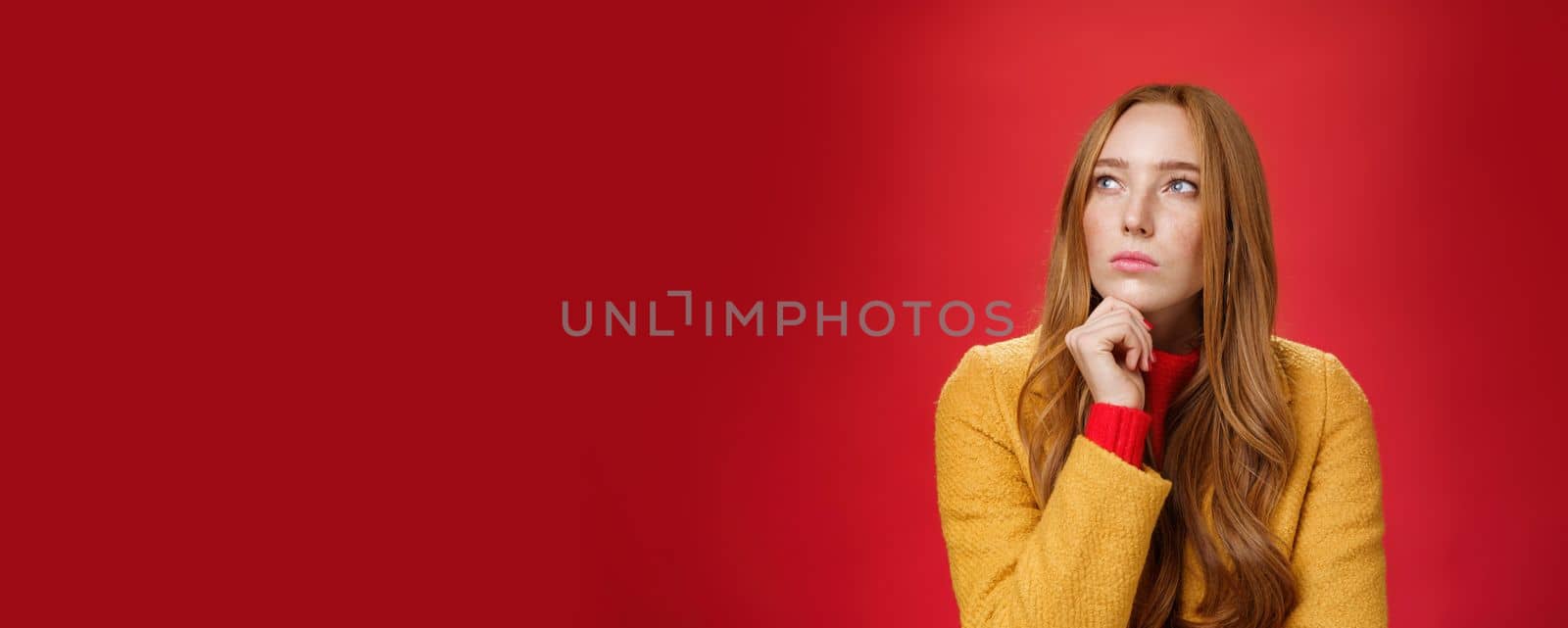 Close-up shot of determined and focused creative thoughtful redhead female looking at upper left corner touching chin, thinking, making choice or remembering information over red background.