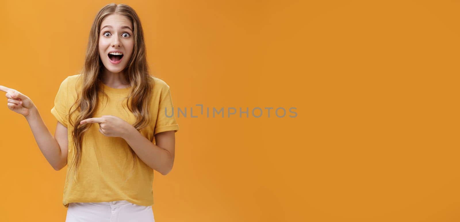 Portrait of excited surprised and amazed cute young european slim woman. with wavy natural hairstyle dropping jaw astonished pointing left asking question about awesome thing over orange wall.