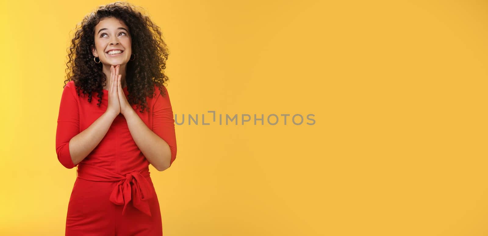 Lifestyle. Dreamy excited cute b-day girl with curly hair in cute red dress rubbing palms together near chest as hands in pray smiling looking up delighted and hopeful making wish over yellow background.