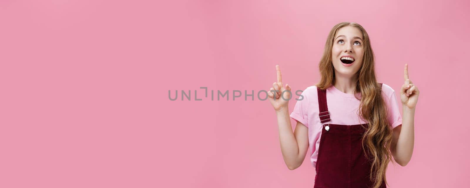 Lifestyle. Amazed and delighted dreamy young teenage girl in stylish corduroy overalls smiling broadly with thrilled smile looking and pointing up joyfully enjoying watching plane flying in sky over pink wall.