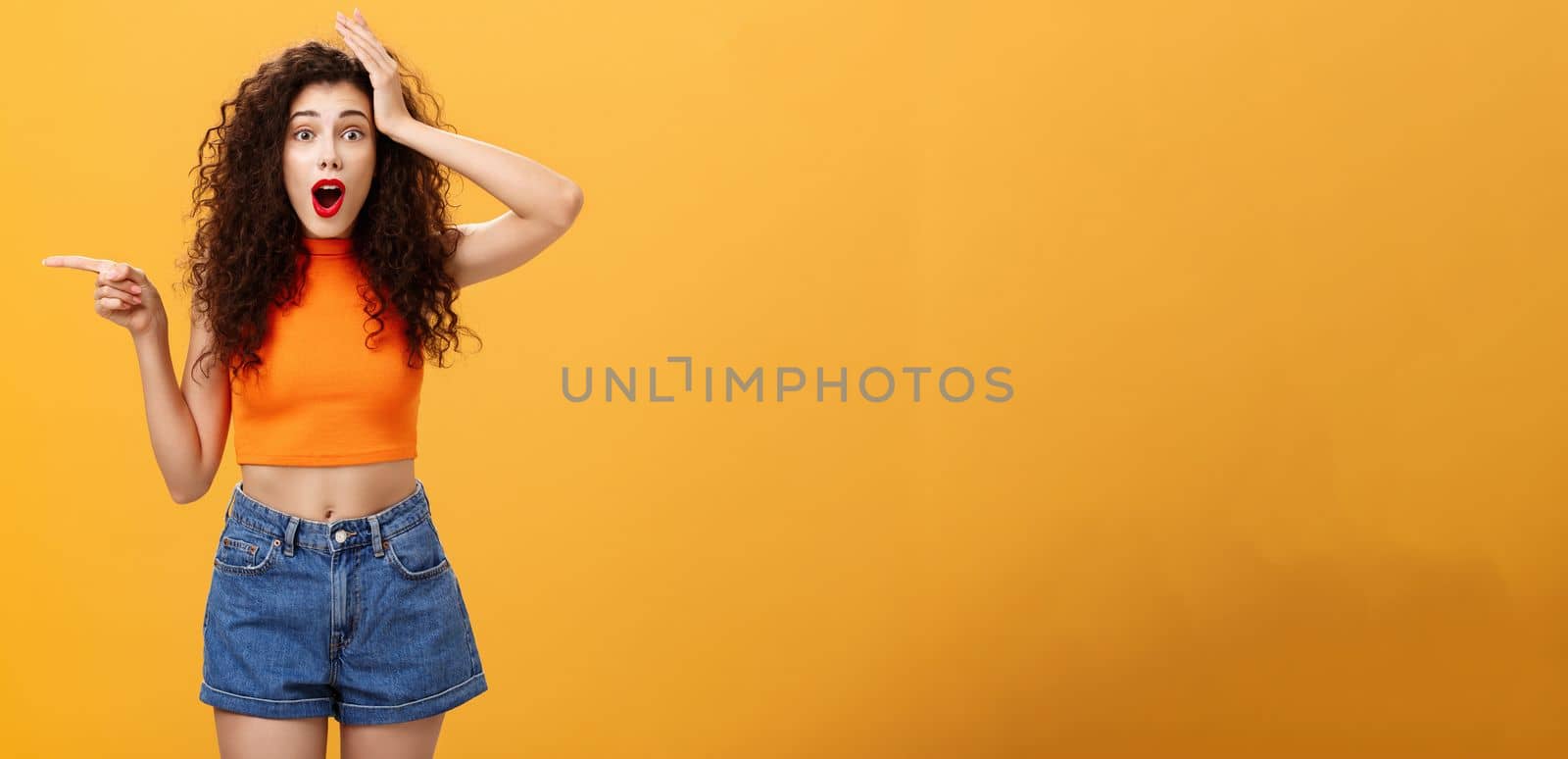 Surprised thrilled beautiful caucasian girl. with curly hairstyle in red lipstic and orange cropped top holding hand on head amazed and pointing left questioned and astonished over studio background.