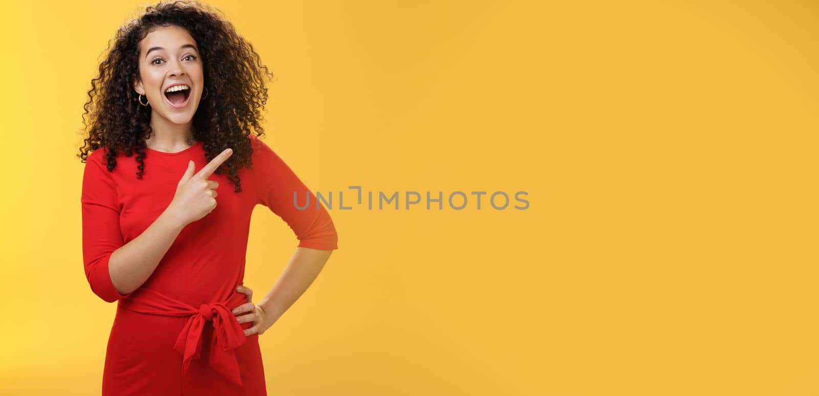 Waist-up shot of amused and impressed beautiful caucasian girl with curly hair in red dress open mouth with amazement and joy and pointing at upper right corner checking out awesome copy space by Benzoix