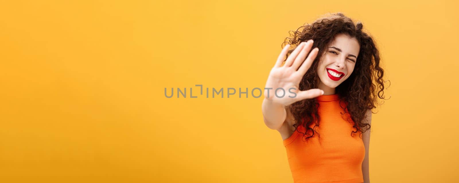 Give me five mate. Stylish friendly and joyful attractive woman with curly hairstyle winking and smiling happily pulling hand towards camera to greet or congratulate friend over orange background by Benzoix