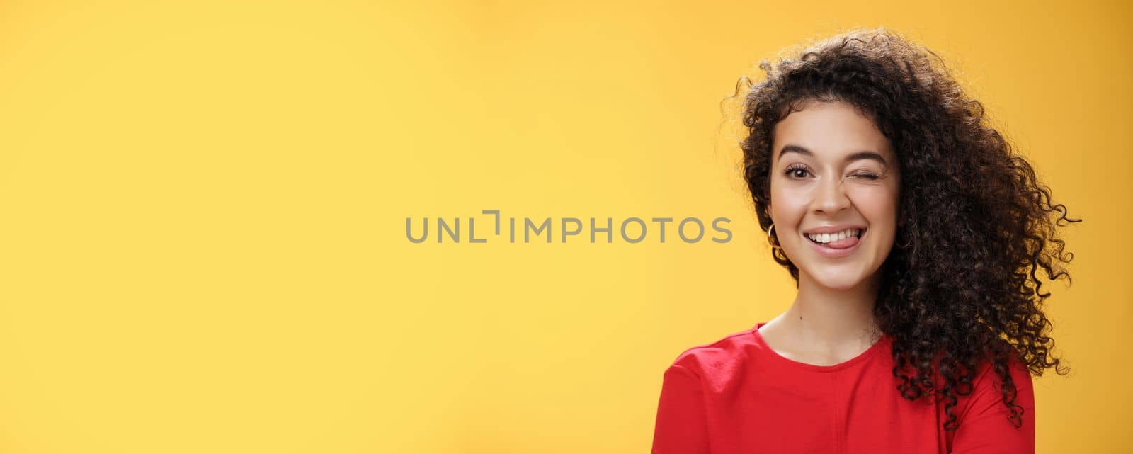 Portrait of funny and cool sister with curly hair winking playfully having fun and foolind around showing tongue as playing with siblings adoring spend time with chidren over yellow background by Benzoix
