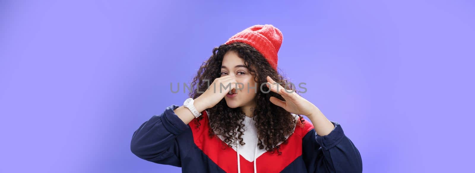 Gosh what stink. Portrait of cute caucasian girl with curly hair in cute trendy hat close nose with fingers and waving hand as smelling awful reek or smell standing disgusted against blue background.