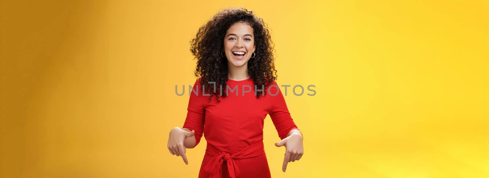 Waist-up shot of assertive charming and happy young woman with curly hairstyle laughing joyfully, smiling and pointing down as showing cool place to check out over yellow background by Benzoix
