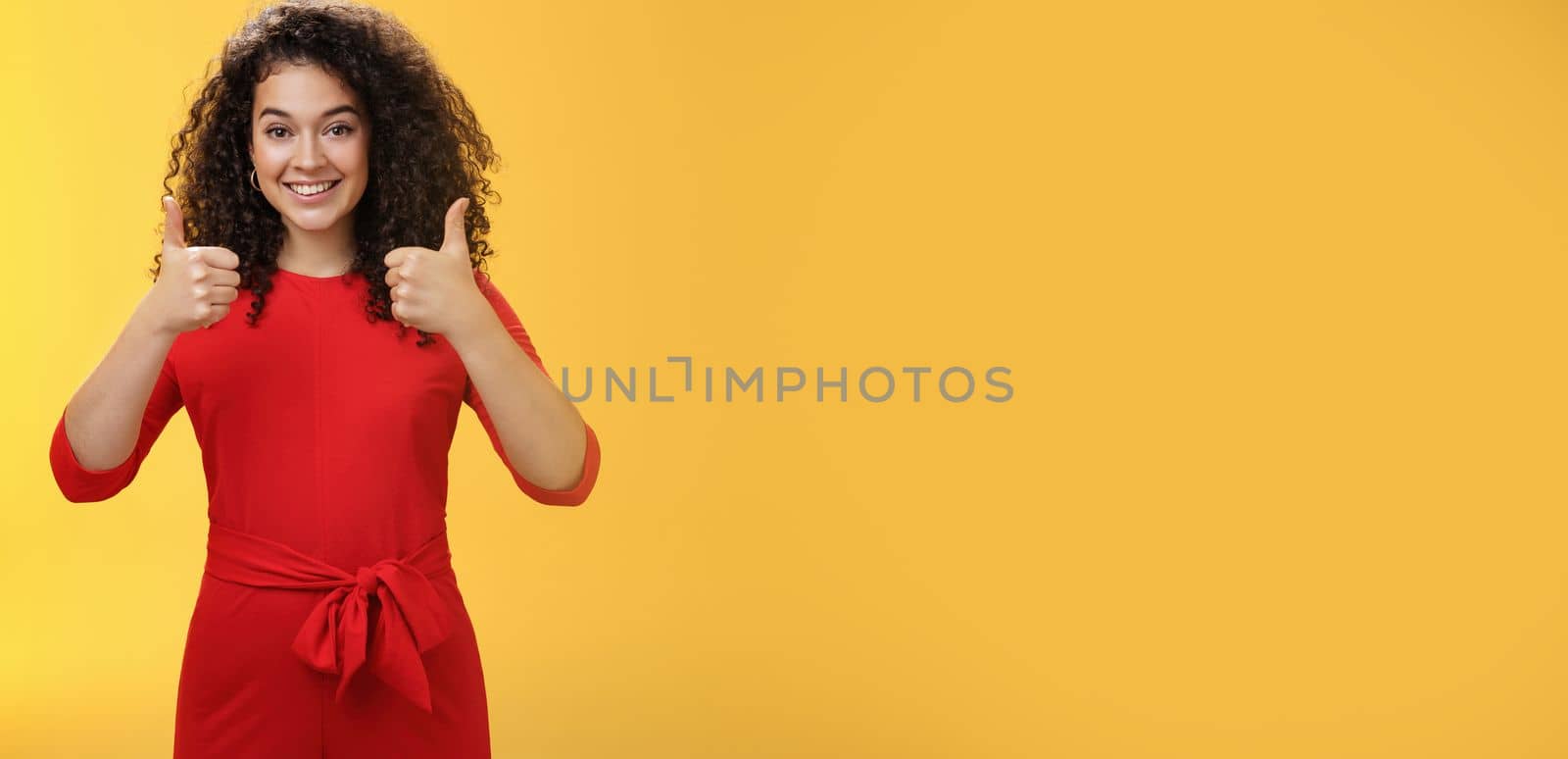 Excellent idea, nice job. Portrait of supportive delighted and happy charming female friend showing thumbs up as standing in red dress over yellow wall smiling giving positive reply, liking concept.