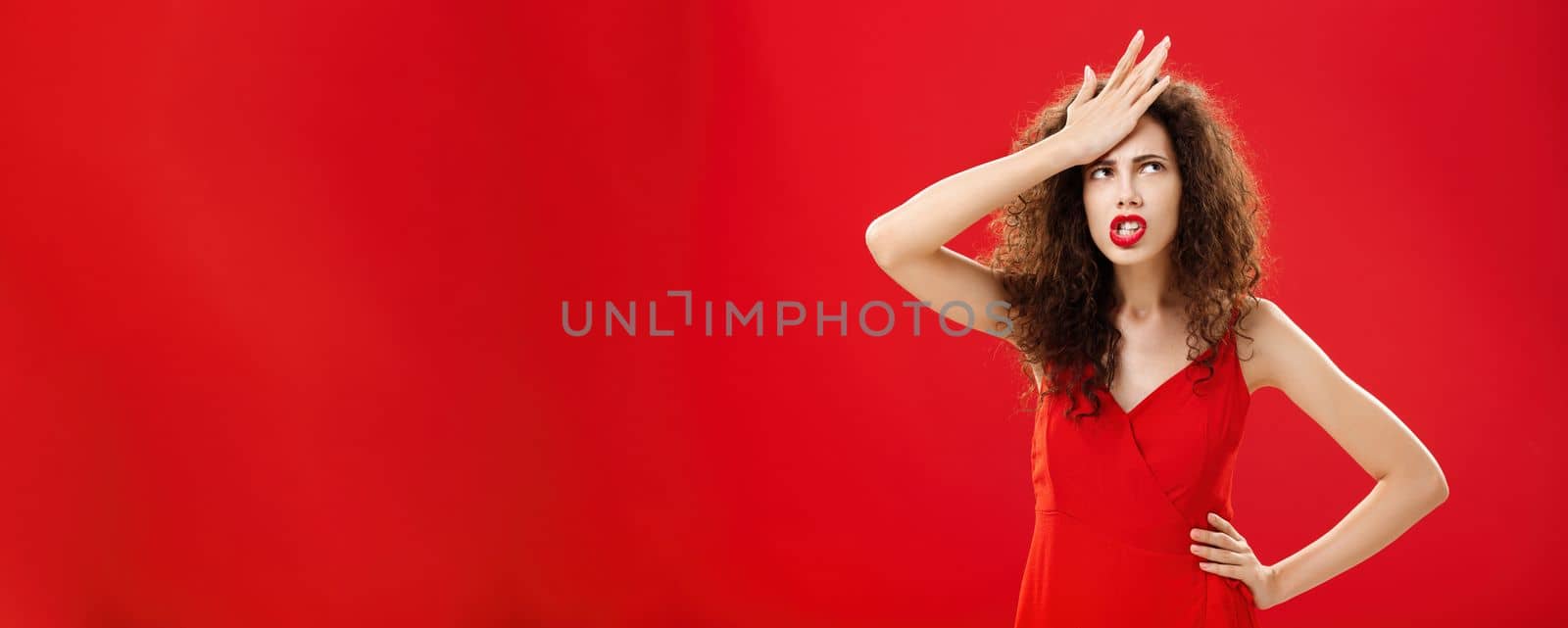 Woman feeling irritated of stupid waiter messing up order punching forehead from annoyance rolling eyes up in anger frowning, swearing saying cursing words displeased standing in red dress. Emotions and body language concept