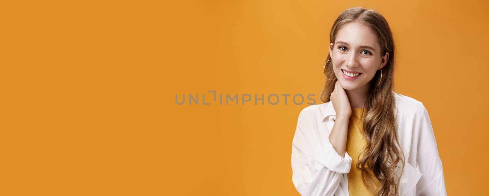 Close-up shot of glamourous young tender girlfriend dressed on date touching neck shy and silly smiling joyfully at camera blushing feeling gentle over orange background in cool outfit by Benzoix