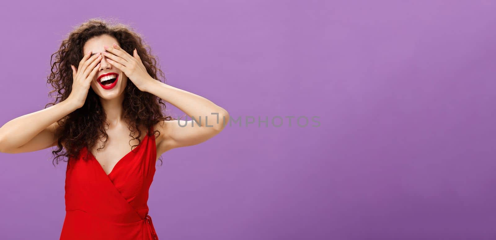 Wife waiting for husband present gift for anniversary closing eyes with palms and smiling broadly peeking through fingers being charmed and excited standing in seductive red dress over purple wall. Emotions concept