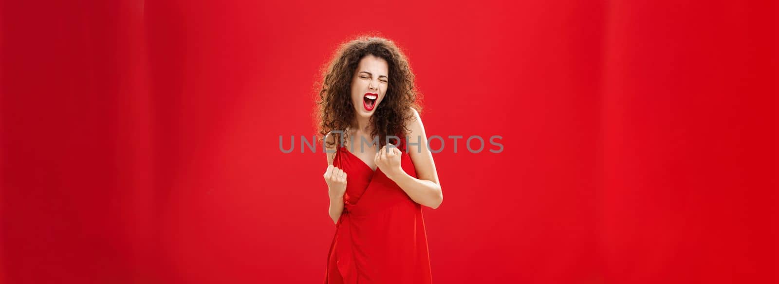 Yeah we are champions. Triumphing happy and delighted rich elegant female with curly hair in stylish dress clenching fists in yes gesture yelling celebrating joyfully win or victory over red wall.