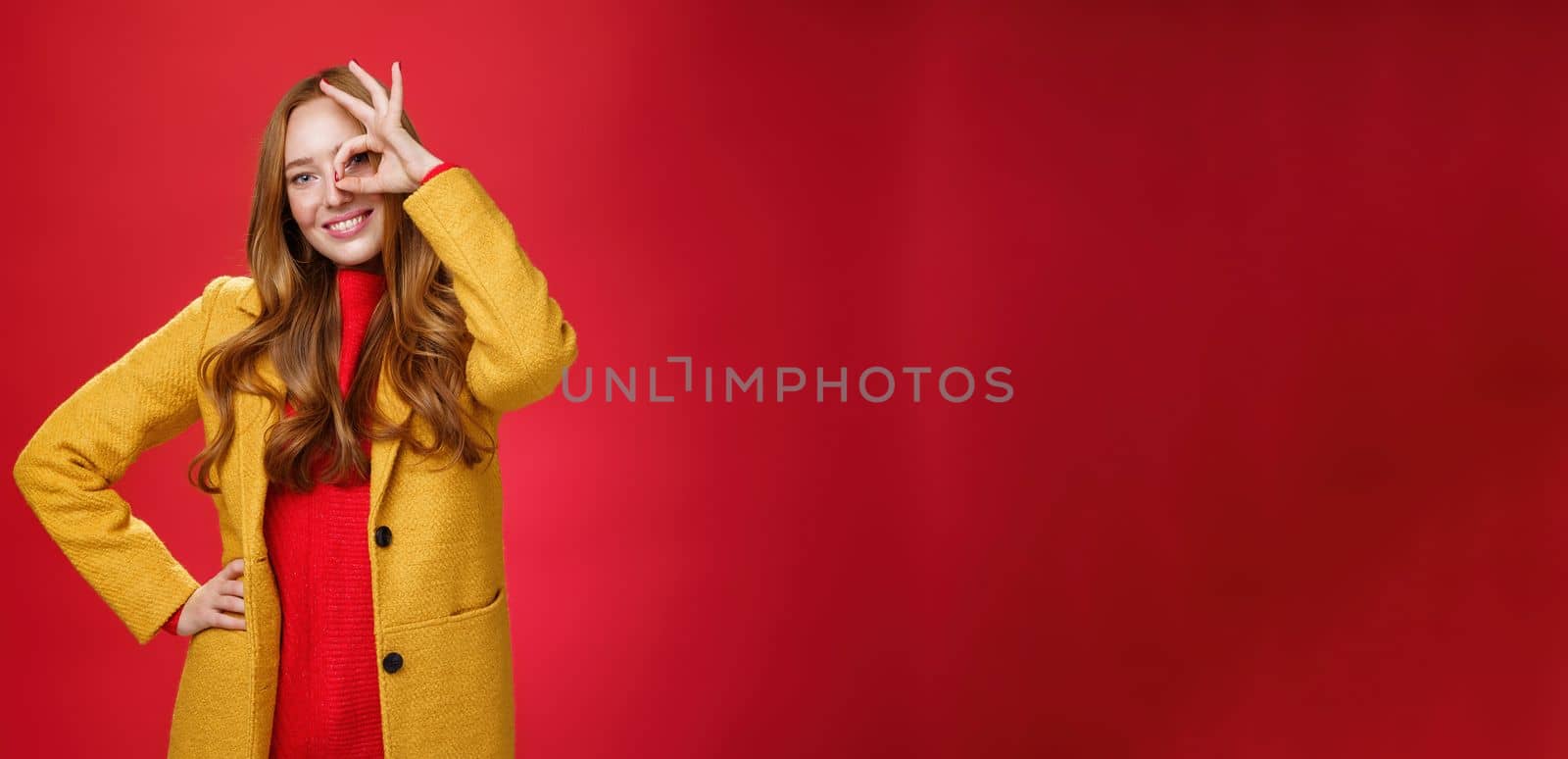 Bright and sunny girl feeling happy even on rainy days standing in yellow stylish coat over red background showing okay gesture over eye, smiling broadly at camera, holding hand on waist.