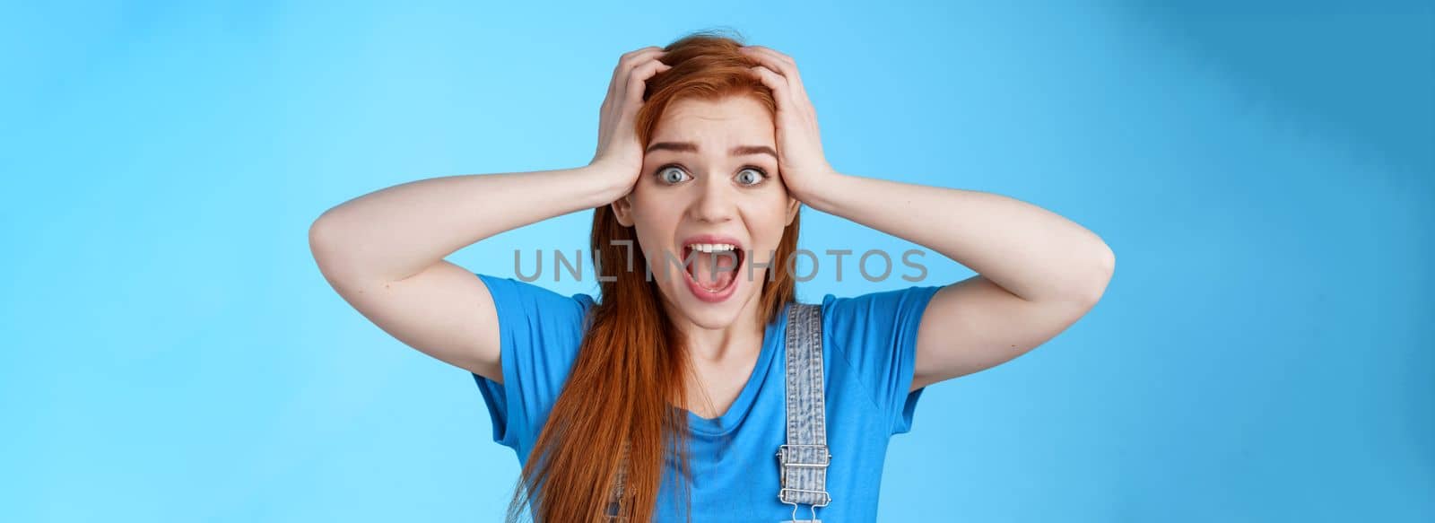 Shocked redhead girl shouting panic, feel pressured deadlines, scared see mess, grab head, scream frightened distressed, stare camera hopeless, anxiously react losing lottery, blue background.