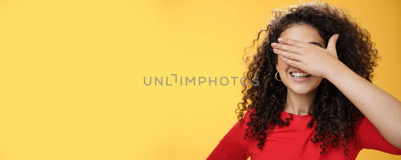 Lifestyle. Close-up shot of dreamy happy young cute woman with curly hair covering eyes with palm as counting or playing peekaboo smiling broadly anticipating surprise happen over yellow background.
