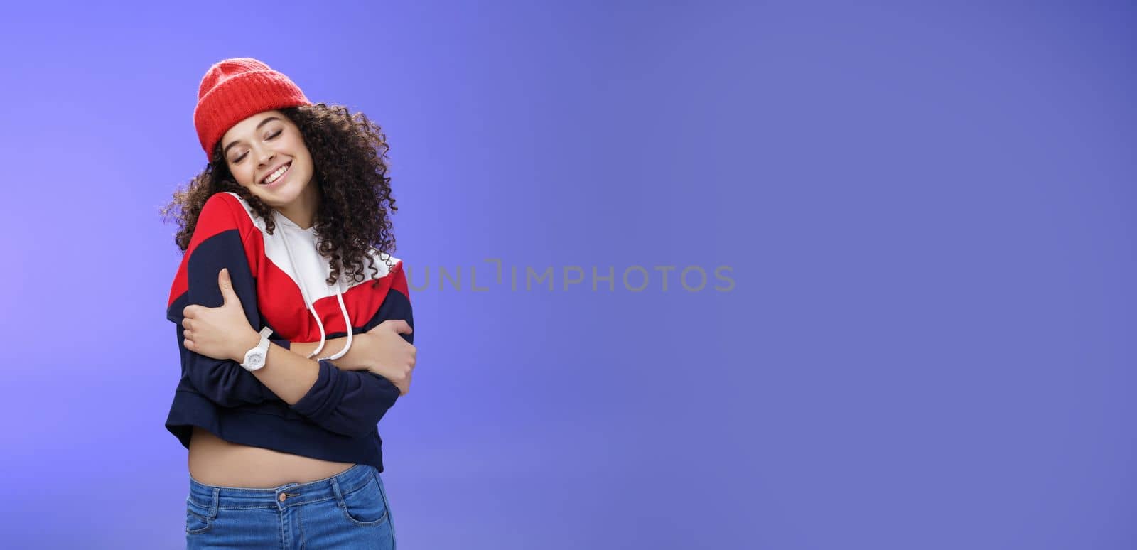 Lifestyle. Sweet tender and sensual curly-haired woman in red stylish beanie hugging herself with closed eyes and nostalgic smile leaning on shoulder feeling warmth and tenderness as recalling loving memories.