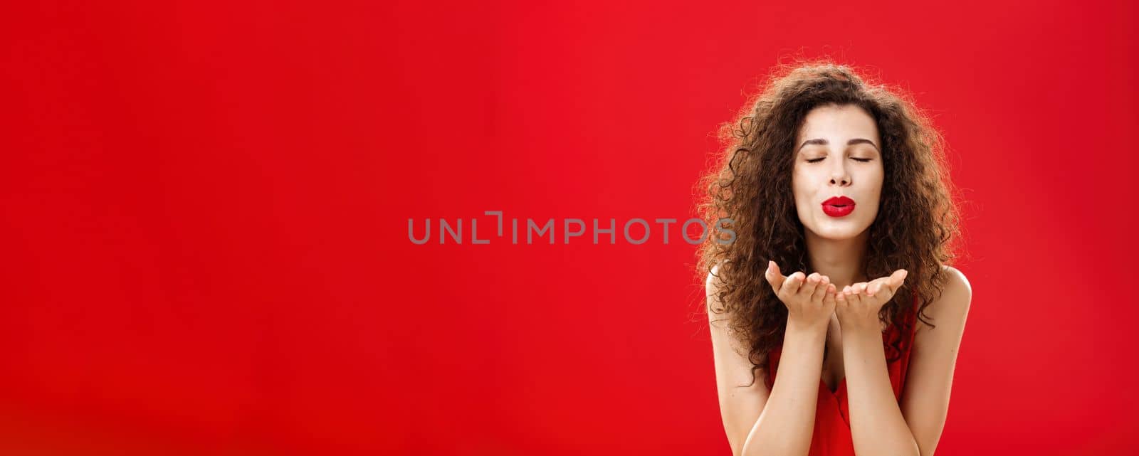 Tender and gentle stylish caucasian girl with curly hairstyle and red lipstick bending towards camera with slight smile closed eyes and palms near folded lips blowing kiss at camera romantically by Benzoix