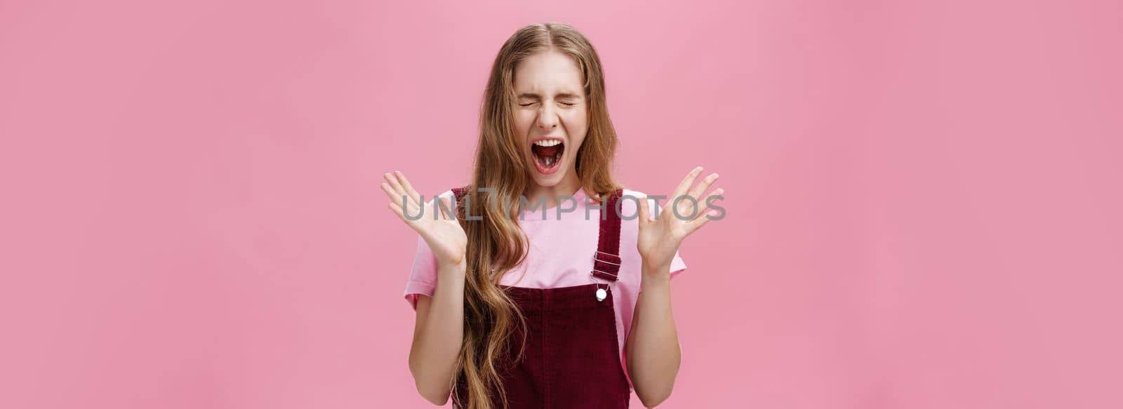 Young girl scared to death seeing spider in room yelling out loud closing eyes and opening mouth gesturing with raised hands near chest feeling afraid and insecure posing against pink background by Benzoix