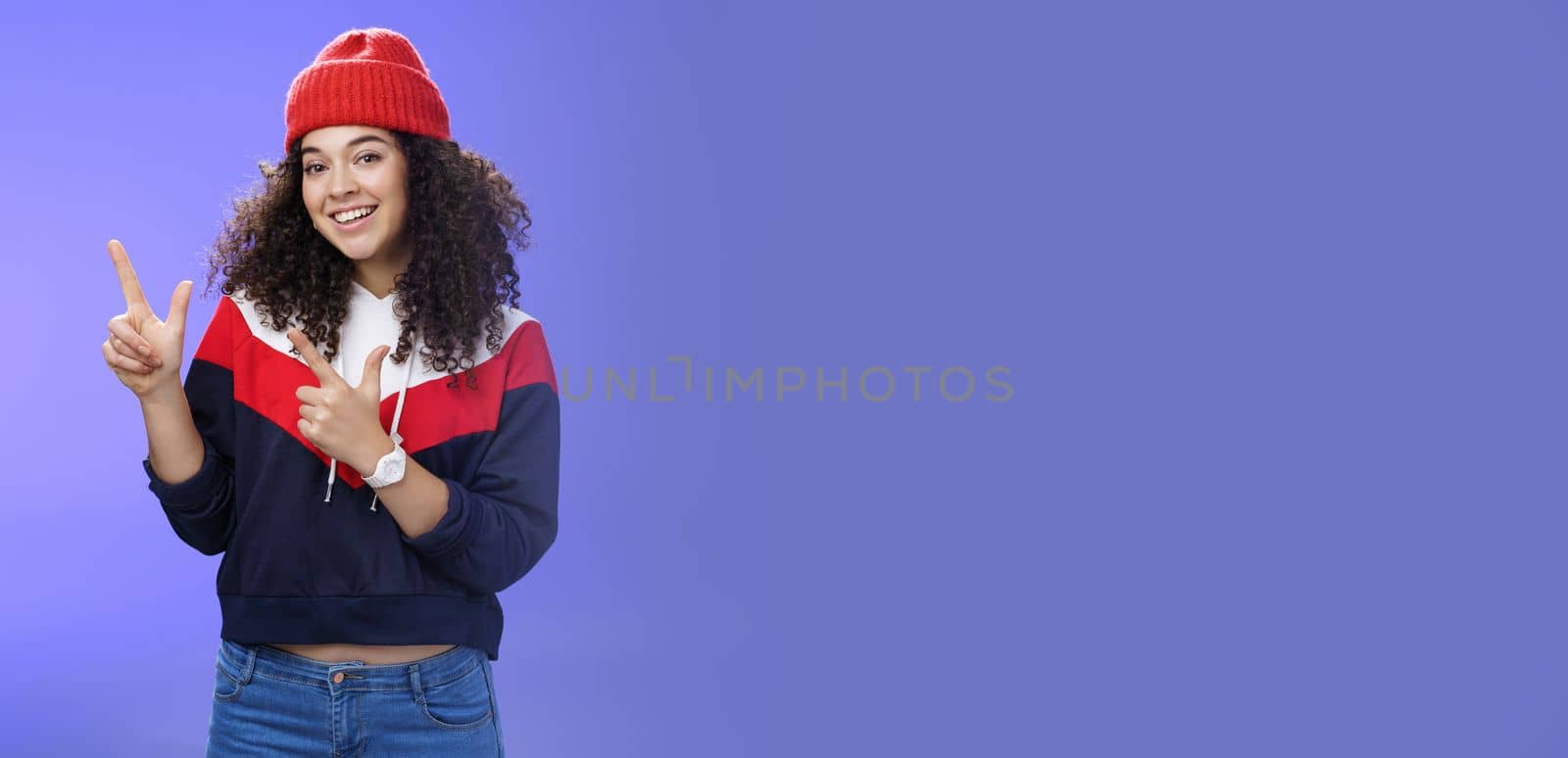 Lifestyle. Portrait of friendly good-looking woman with curly hair in red warm beanie pointing at upper left corner and smiling delighted as promoting cool copy space against blue background.