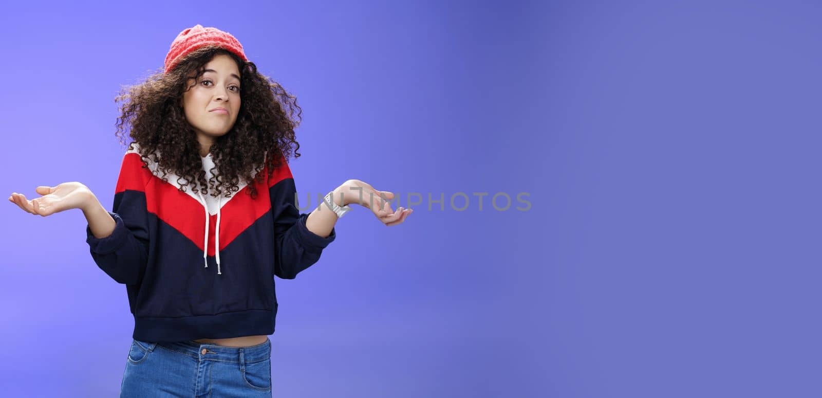 Upset girl cannot get clue what happened shrugging turning away as pouting feeling questioned and unsure what answer, standing clueless and puzzled in sweatshirt and stylish hat over blue background.
