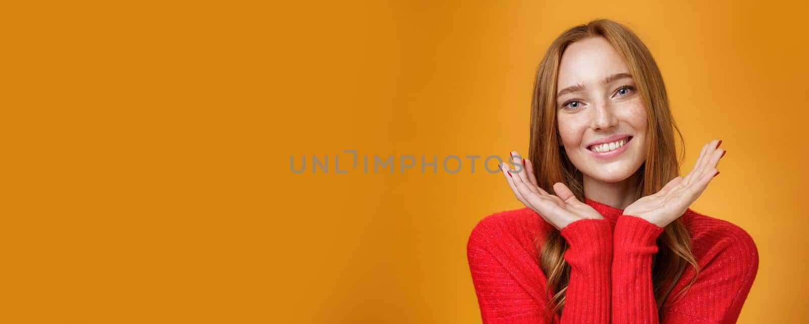 Close-up shot of sensual tender and gentle ginger girl with romance and happy gaze holding palms near pure clean skin with freckles smiling joyfully tilting head looking friendly over orange wall by Benzoix