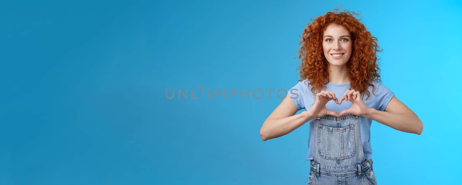 Tenderness, love, romance concept. Lovely feminine cute redhead curly woman show heart gesture chest express sympathy passionate feelings smiling broadly cherish feelings standing blue background by Benzoix