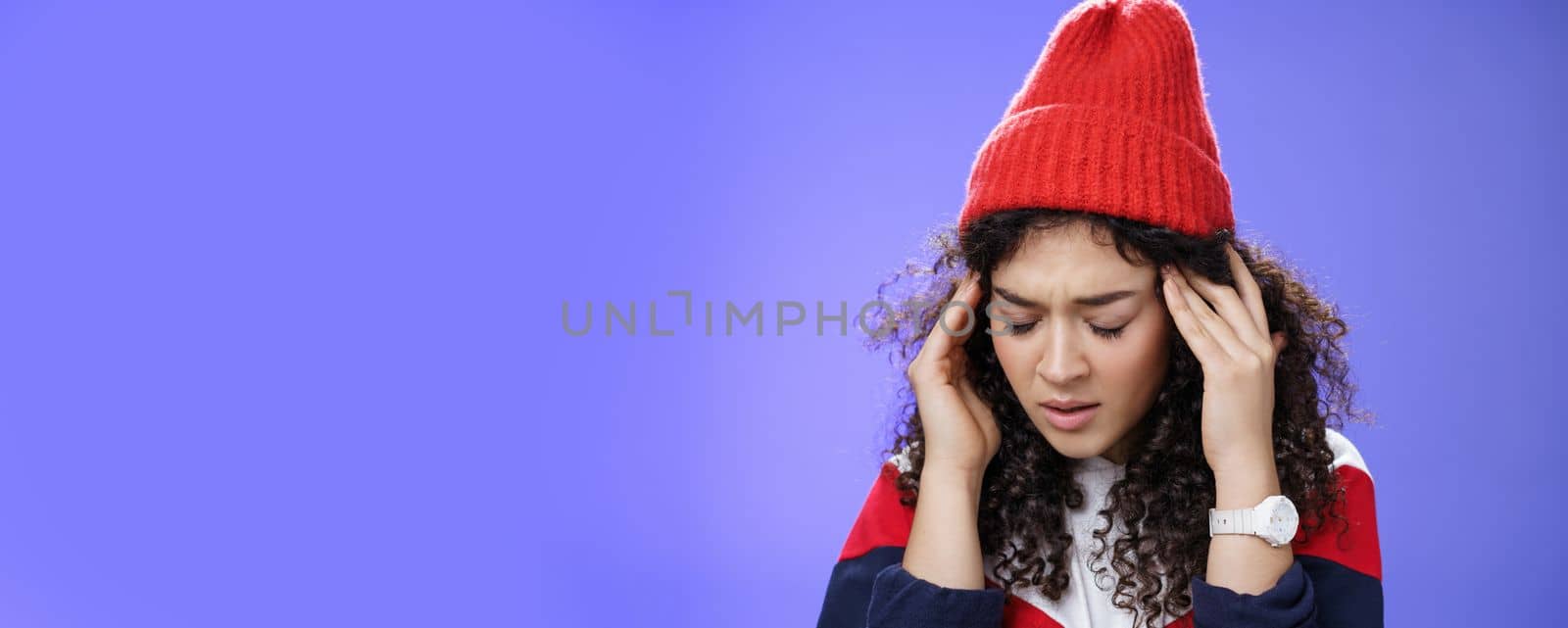 Close-up shot of sad and sick cute stylish woman in red warm beanie close eyes rubbing temples feeling tired and suffering headache as catching cold, standing with terrible migraine over blue wall.