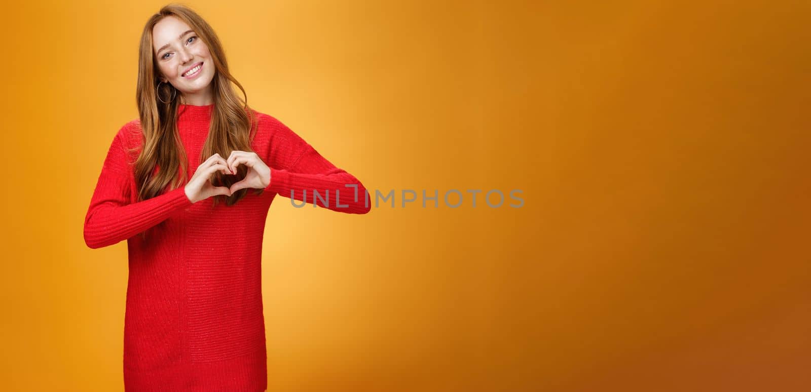 Lifestyle. Attractive confident and cute ginger girl with freckles in red knitted warm dress showing love gesture tilting head and smiling broadly at camera liking and adoring new outfit over orange background.