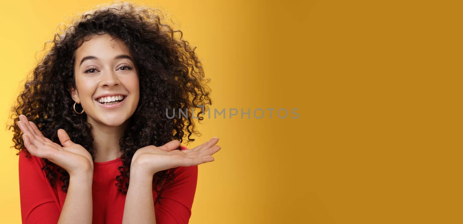 Lifestyle. Close-up shot of happy kind and tender pretty caucasian female student with curly hair and perfect skin smiling delighted holding palms spread near face having fun over yellow background.