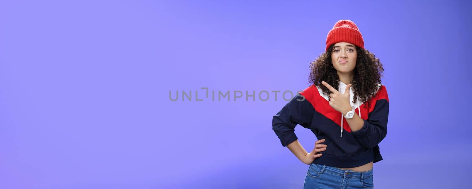 Girl feleing sad missing good chance, such shame. Portrait of displeased woman regretting frowning and pursing lips gloomy pointing at upper left corner upset wearing warm beanie over blue wall by Benzoix