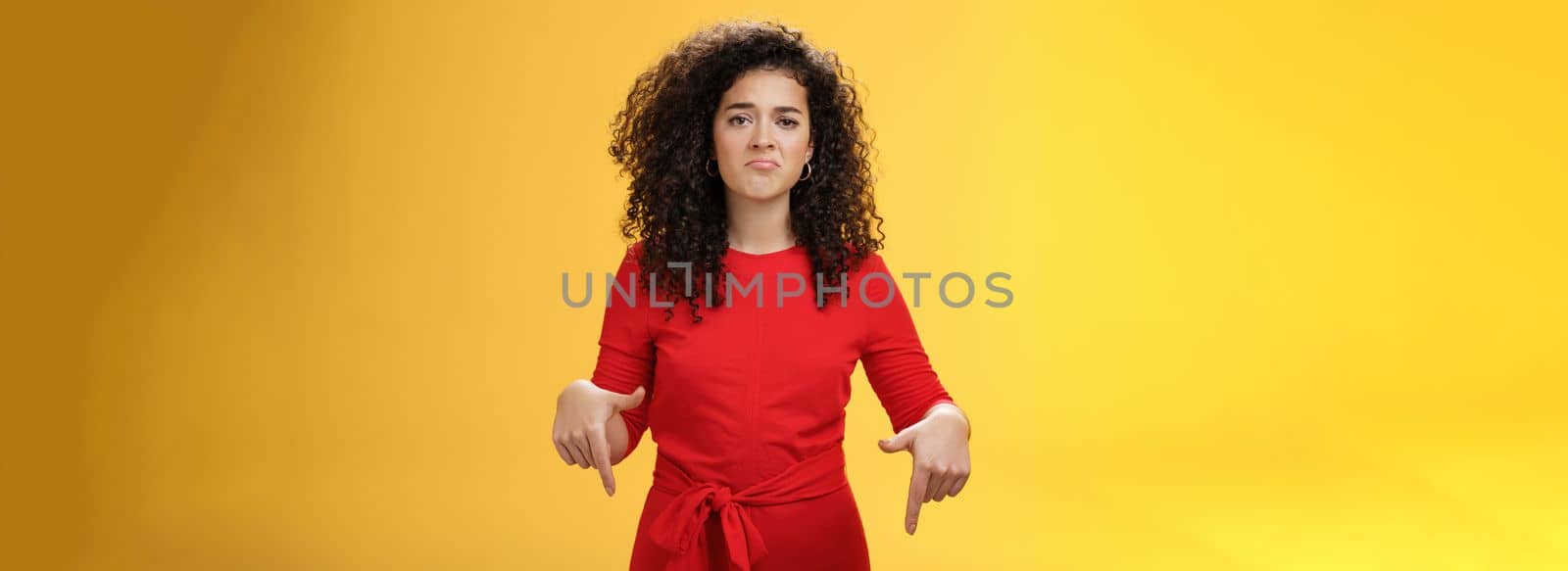 Portrait of gloomy girl with sad face pursing lips down frowning disappointed and pointing down with regret and unsatisfied expression, missing opportunity feeling upset over yellow background.