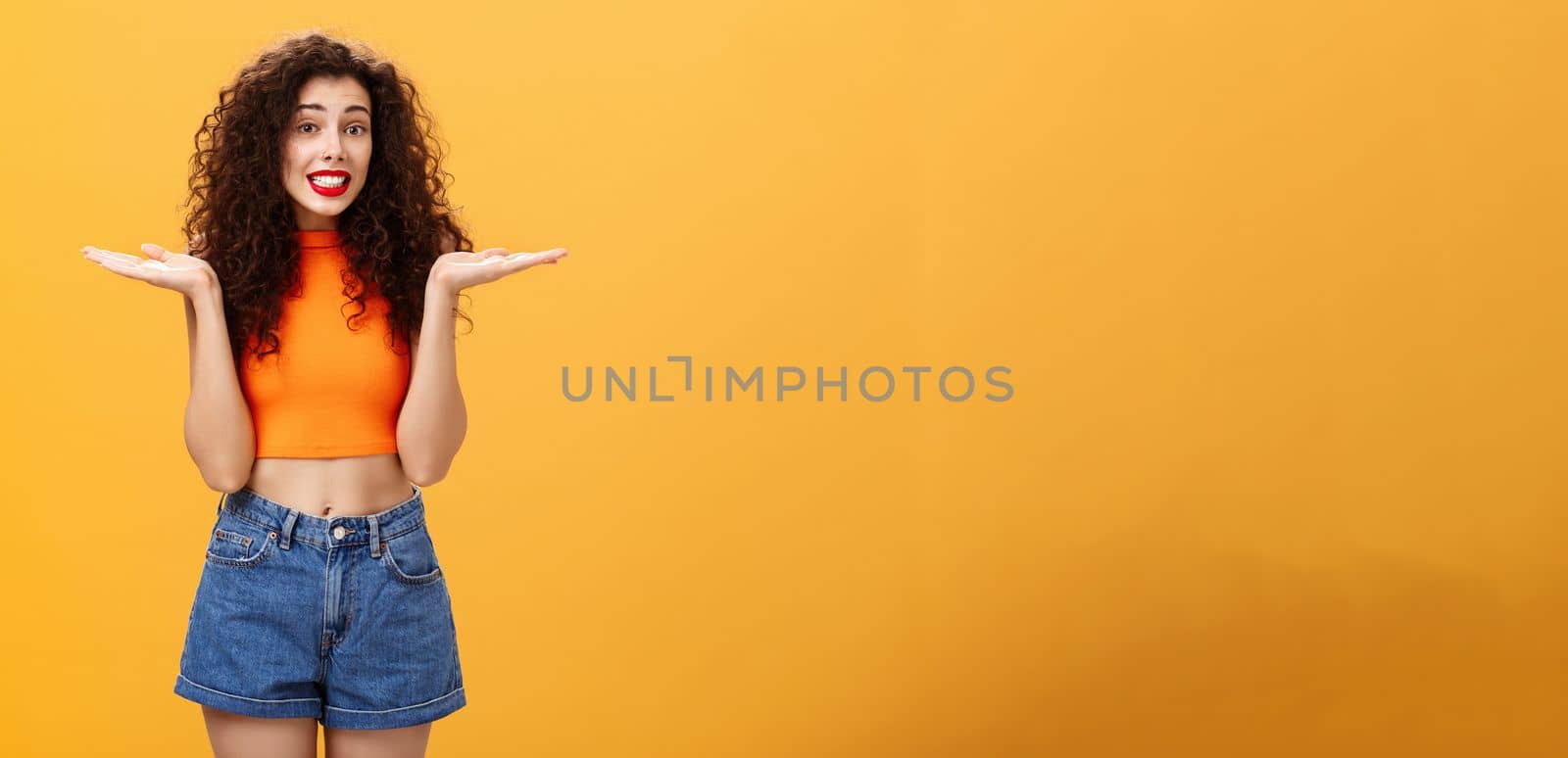 Oops honey sorry. Clueless silly stylish european female with curly hairstyle in cropped top shrugging with hands spread aside in unaware gesture smiling guilty and questioned over orange background by Benzoix