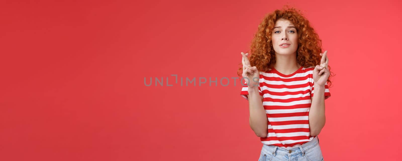 Lifestyle. Nervous young silly timid cute redhead ginger girl anticipating hopeful results believe praying squinting intense worry to win cross fingers good luck wish come true red background.