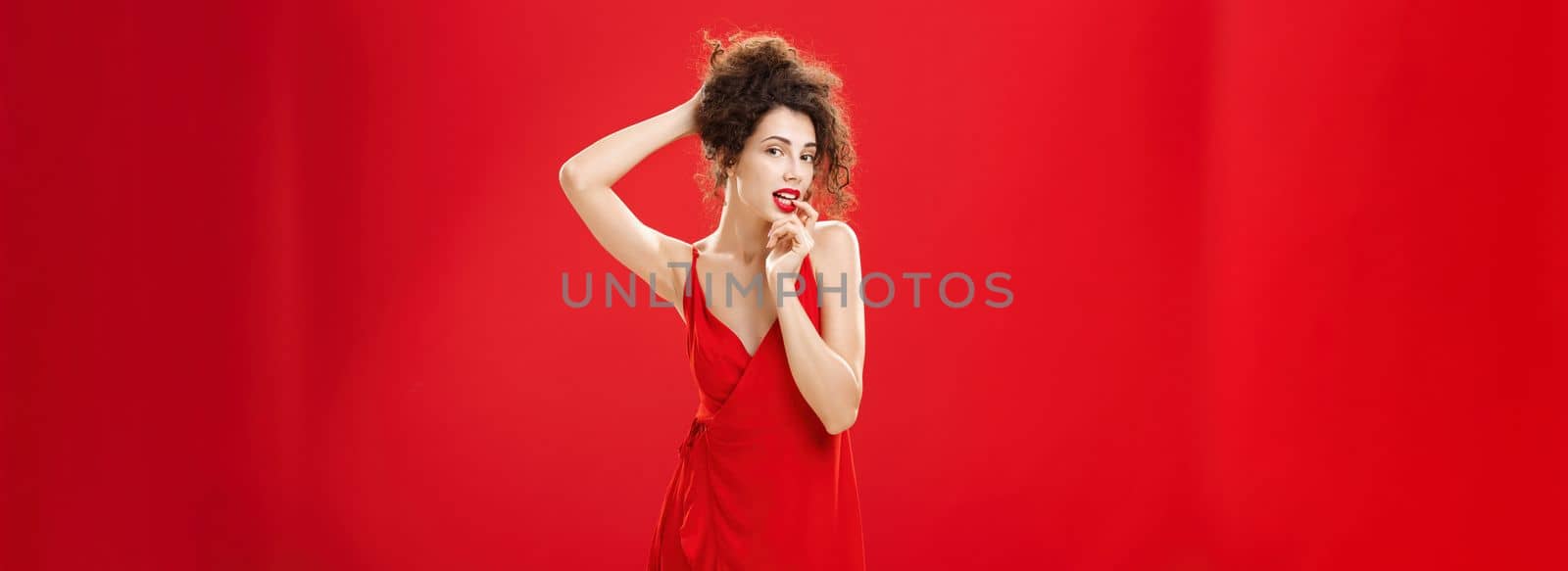 Seductive and sensual elegant rich lady in red evening dress combing hair in luxurious hairstyle touching lip flirty standing over studio background forplaying with husband after romantic dinner. Copy space