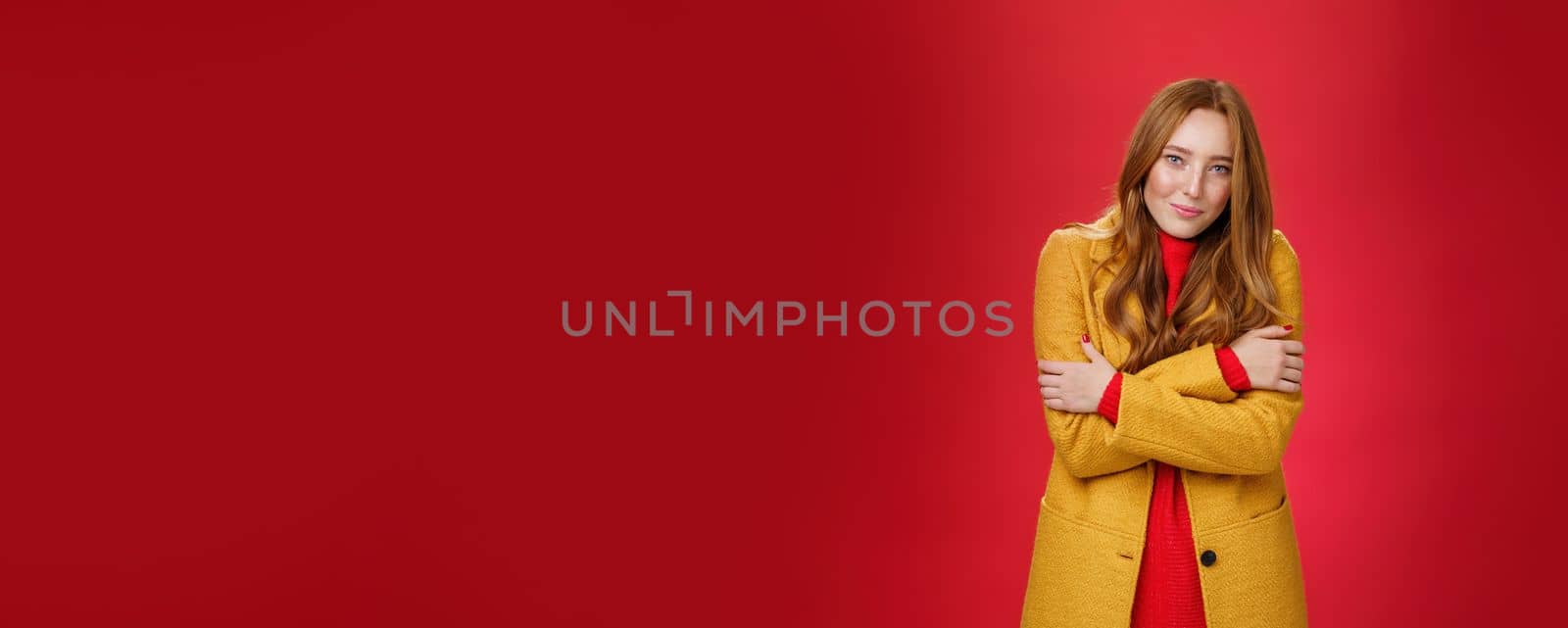 Romantic good-looking tender and cute ginger girlfriend with freckles hugging herself and looking with sensual gentle smile at camera getting cold or chilly waiting outside over red background by Benzoix