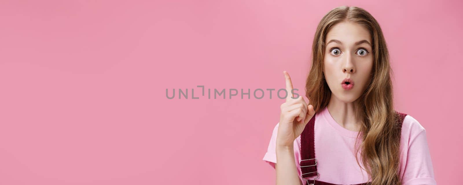 Close-up shot of creative emotive young cute female student with beautiful natural wavy hair raising index finger in eureka gesture folding lips while saying suggestion amazed and thrilled by Benzoix