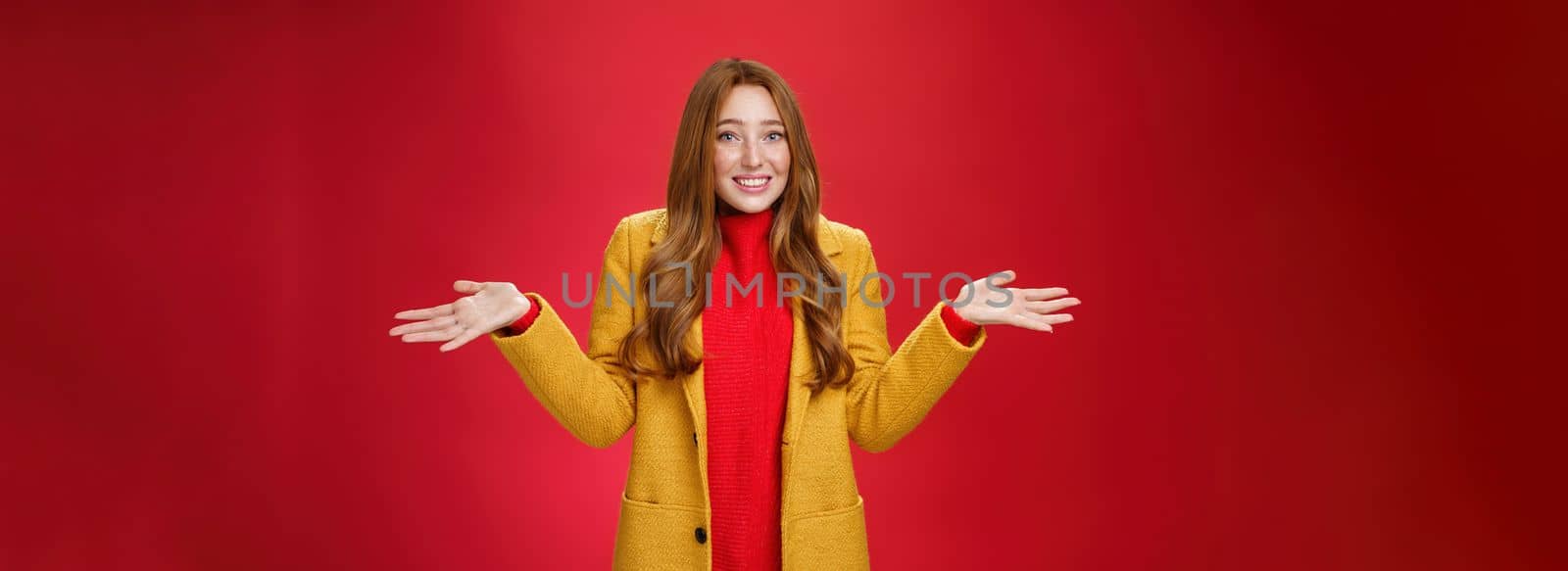 Redhead girl apologizes for being late smiling silly and guilty. with sorry look shrugging as raising hands sideways wearing stylish yellow coat over dress posing against red background.