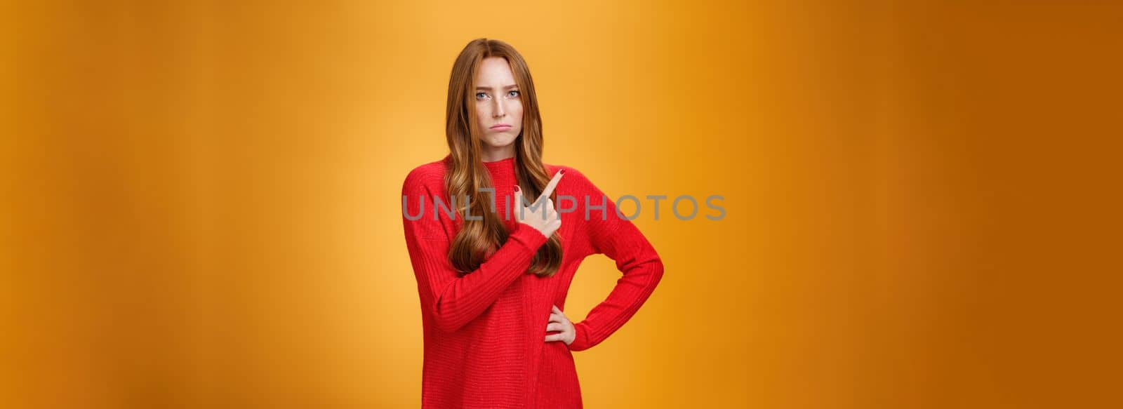 Upset and disappointed young ginger girl in red sweater pointing at upper left corner or behind frowning looking gloomy and angry, standing jealous and moody against orange background by Benzoix
