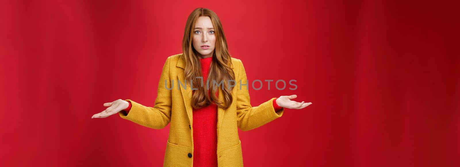 Upset and confused redhead girlfriend cannot understand what happened shrugging with spread hands sideways and clueless grimace, being unaware why, posing concerned over red wall by Benzoix