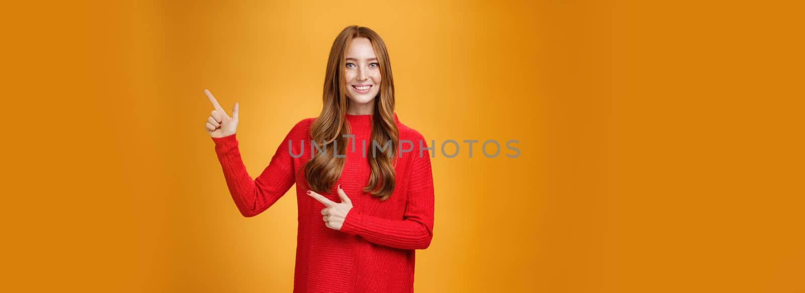 Friendly-looking joyfuly and energized redhead female in red sweater pointing at upper right corner promoting advertisement with broad delighted and tender smile posing against orange background by Benzoix
